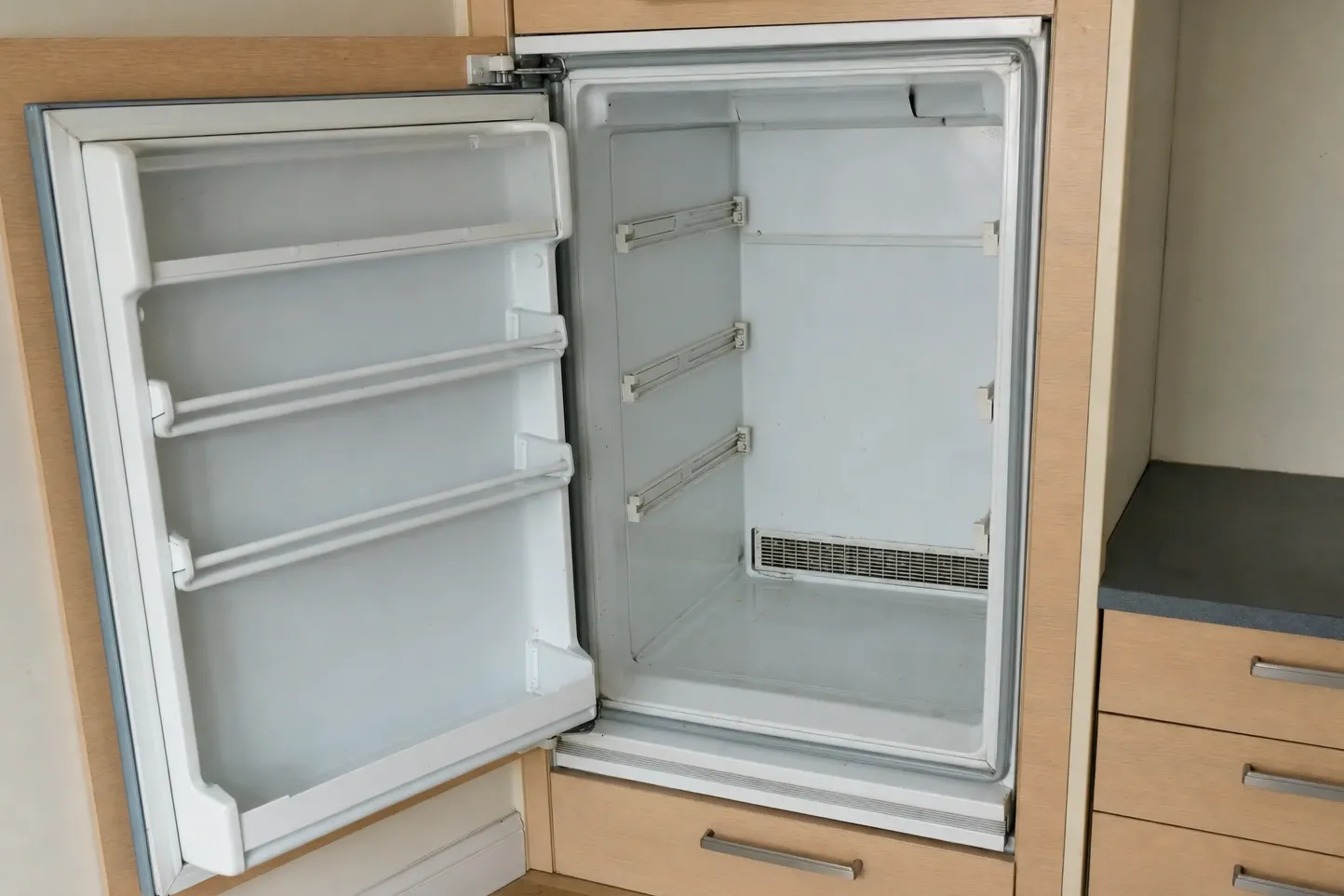 Older undercounter refrigerator built into light wood cabinetry, door open on empty shelves and an interior vent grille